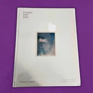 Front view of the 'Imagine John Yoko' hardback book. The cover is minimalist white with a centered Polaroid photo of John Lennon, overlaid with transparent clouds. The book's cellophane wrapping reflects light and shows minor wear but remains sealed.